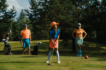 Obraz premium A young girl prepares to hit a ball on a sunny golf course surrounded by her parents, playing a family game of golf on a weekend morning