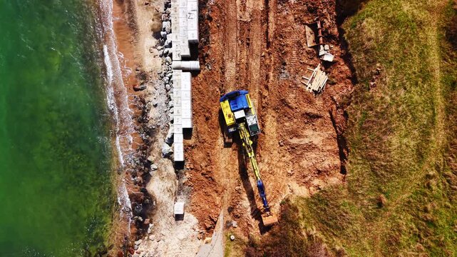 Excavator building coastal seawall on Black Sea shore. Overhead drone shot of concrete blocks, red soil and turquoise water along the beach.