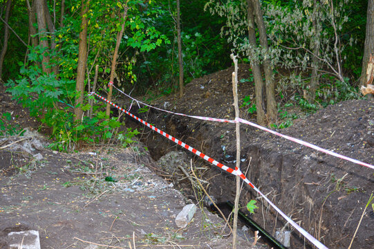 Long excavated ground trench in a forest area cordoned off with red and white safety caution tape. Outdoor pipeline installation, earthworks, and utility excavation safety concept.