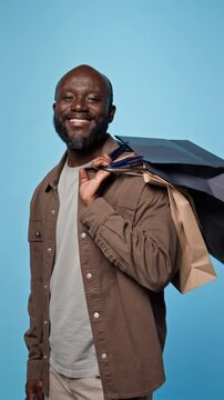 Portrait of African American man placing shopping bags on shoulder and smiling. Visual fits retail marketing, holiday sale campaigns and commercial ecommerce promotions, vertical