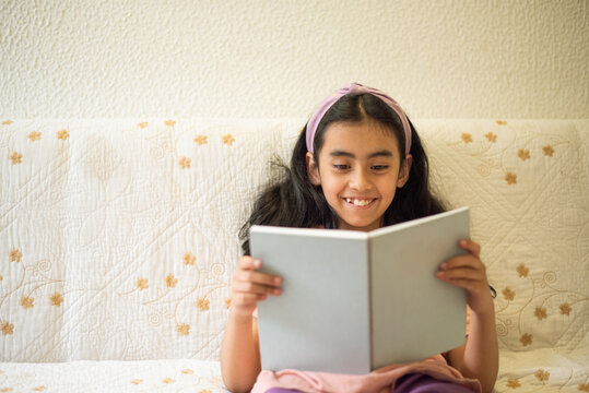 Smiling girl reading a book at home while sitting on a sofa, portraying childhood education, literacy, curiosity and relaxed independent learning.