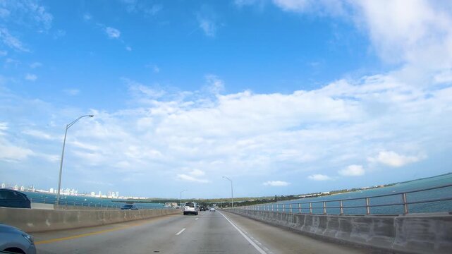 Driving across a bridge toward Miami Beach under a clear blue sky, capturing the urban coastal landscape and atmosphere of South Florida.