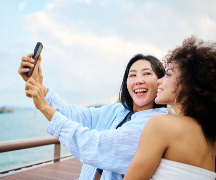 Two friends laugh and pose for a selfie on a waterfront boardwalk overlooking the sea. The smiling women enjoy a relaxed seaside outing, capturing travel memories with a smartphone against a backdrop 