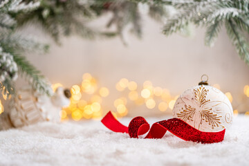 Naklejka premium A Christmas background featuring an elegant white and gold patterned bauble with a curled red glitter ribbon beneath frosted pine on snow, softly lit by warm twinkling lights with copy space.