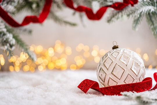 A Christmas background featuring a white bauble with an etched diamond pattern beside a sparkling red ribbon beneath frosted pine on snow, softly lit by warm twinkling lights with copy space.