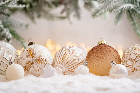A Christmas background featuring a row of textured white and gold baubles with etched patterns beside a bronze glitter ornament beneath frosted pine on snow, softly lit by warm twinkling lights.
