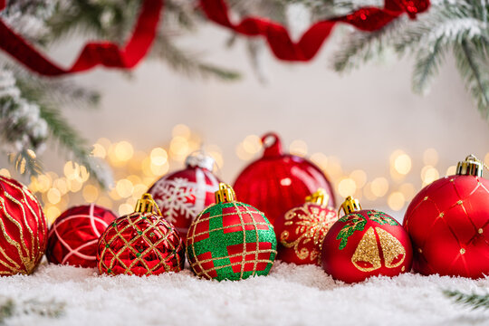 A Christmas background featuring a row of red baubles with varied gold glitter patterns, bells, and holly beneath frosted pine on snow, softly lit by warm twinkling lights with copy space.