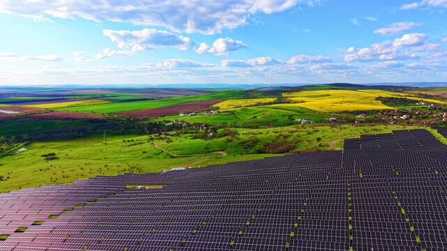 Aerial view of solar farm among rural fields. Panel rows sit beside patchwork farmland, village houses and yellow rapeseed fields under cloudy sky.