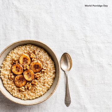A ceramic bowl of oatmeal garnished with caramelised banana coins, walnuts and a sprinkle of cinnamon above beside a vintage silver spoon on a white linen surface.