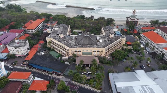 Aerial drone view of Sunrise Hotel in Seminyak, Bali, showing a beachfront resort complex, nearby shops, palm trees and coastal streets at sunrise with tropical urban beachfront atmosphere