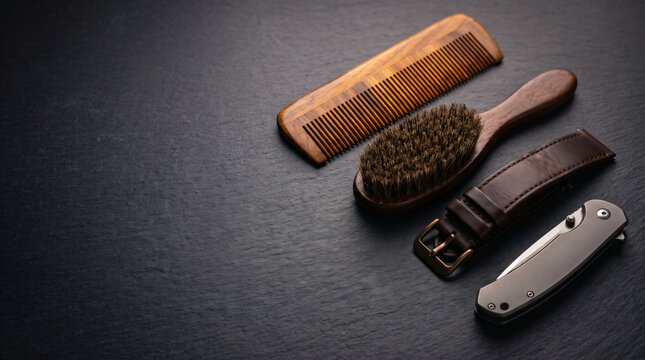 Wooden grooming comb, hair brush, leather watch strap, and pocket knife arranged on dark slate background