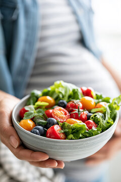 Close up of a pregnant woman holding a bowl filled with fresh salad, cherry tomatoes, and blueberries.
