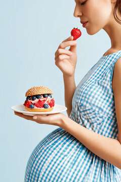 Pregnant woman holding a sweet fruit burger filled with berries and eating a fresh strawberry.