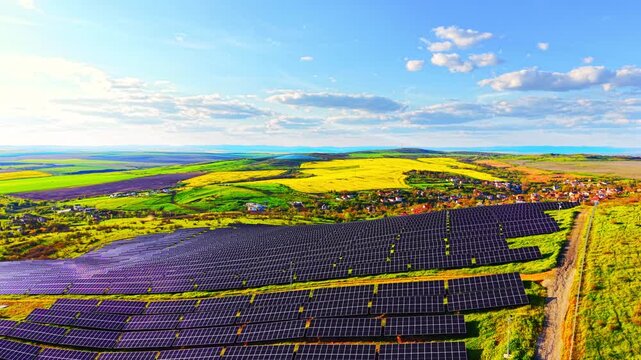 Panoramic solar farm bordering yellow rapeseed field. Wide view shows village rooftops, green meadow and distant hills under blue sky.