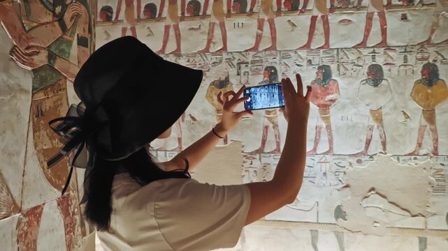 A beautiful tourist wanders inside the tomb of King Seti I, wearing a black hat, holding her mobile phone, and takes pictures of the inscriptions and drawings carved on the walls of the tomb.