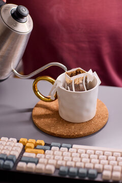 Overhead view of white ceramic coffee mug with paper pour-over dripper, placed on cork coaster beside mechanical keyboard and stainless gooseneck kettle on desk workspace