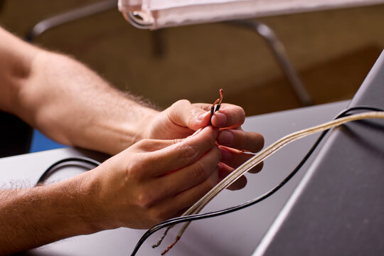 Two hands holding stripped copper wire ends beside electronic enclosure during wiring repair, warm studio lighting with closeup detail of exposed conductor strands