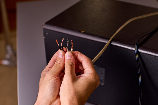Two hands holding stripped copper wire ends beside electronic enclosure during wiring repair, warm studio lighting with closeup detail of exposed conductor strands