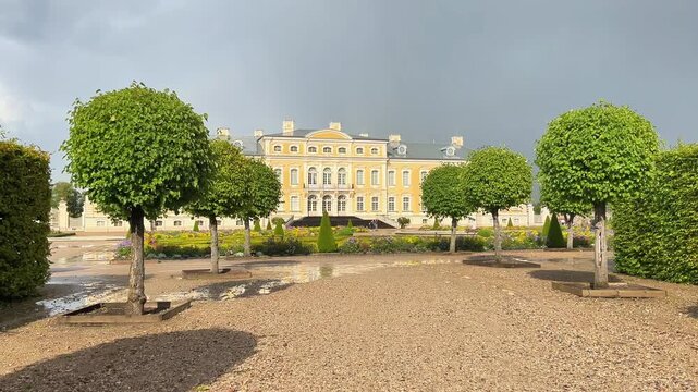 Wide shot of Rundale Palace in Latvia featuring manicured French garden trees and gravel courtyard under a dramatic stormy sky. Travel landmark concept.