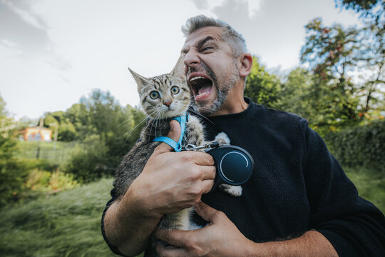 Middle-aged man with gray hair holds a surprised cat on a leash outdoors, expressing excitement in a green park with trees and grass in the background