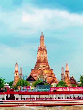 Wat Arun temple standing beside the Chao Phraya River in Bangkok, Thailand, with a traditional long-tail boat crossing the water under a bright cloudy sky, creating a scenic travel view.