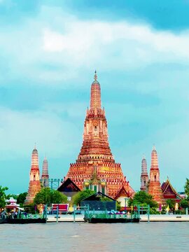 Wat Arun temple standing beside the Chao Phraya River in Bangkok, Thailand, with a traditional long-tail boat crossing the water under a bright cloudy sky, creating a scenic travel view.