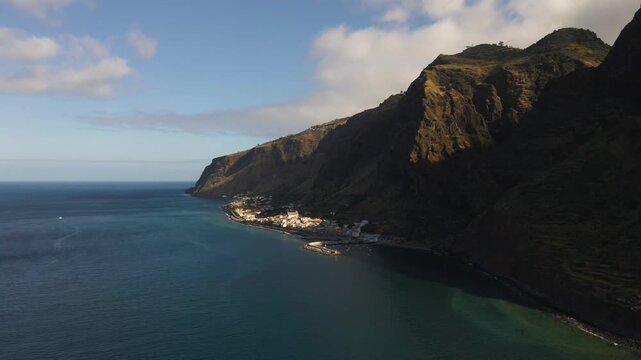 Drone footage capturing the raw natural beauty of Madeira&rsquo;s southern coastline, focusing on Jardim do Mar. The camera flies parallel to the cliffs, revealing the layered terrain, textured rock faces.