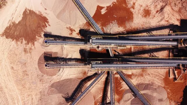 Elevated view of twin conveyor belts over sand pile. Symmetrical drone image of parallel metal conveyors casting shadow on reddish brown quarry ground.