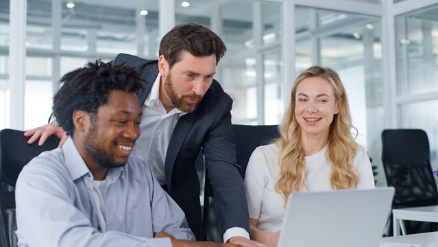 Three multiracial business team in office Caucasian leader boss talking happy satisfied with online laptop good result achieve talk to colleagues coworkers multiethnic group teamwork job with computer
