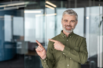 Mature businessman with a confident smile pointing two fingers to the left, indicating a clear copy space against a modern office background with glass walls