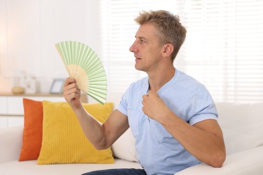 Man waving hand fan to cool himself on sofa indoors