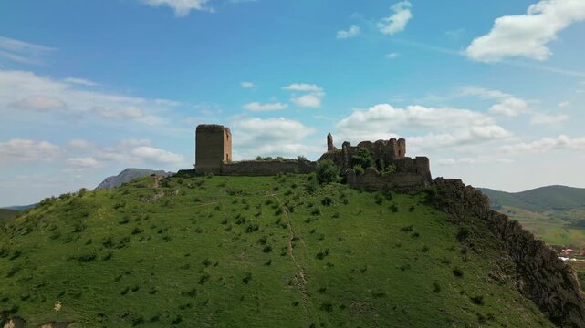Medieval fortress ruins standing on a rocky mountain slope with green hills and blue sky