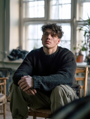 Obraz premium Thoughtful young man with curly hair wearing dark knitted sweater sitting indoors on chair near window, deep in thought, portrait representing solitude, reflection, and quiet contemplation mood