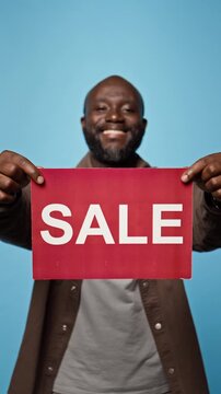Portrait of smiling African American man showing sale sign against blue background. Ideal footage for retail promotions, seasonal shopping discounts, e-commerce marketing, vertical