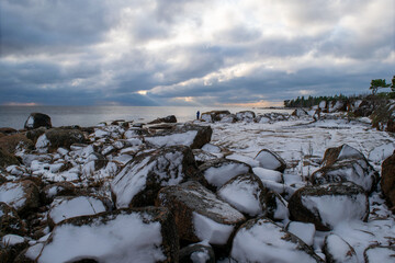 Winter coastal landscape with snow‑covered rocks and a calm shoreline under dramatic cloudy light. A lone person in blue stands near the water, adding scale to the rugged scenery framed by trees and r © Julius