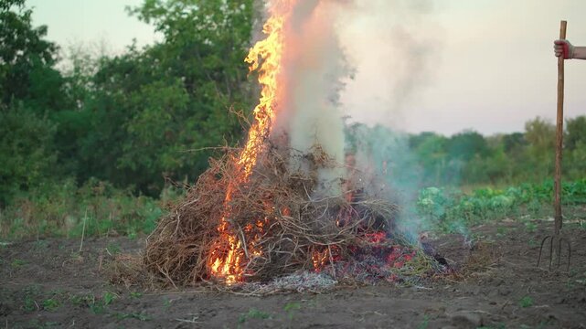 Gardener in gray overalls uses a pitchfork to tend to a burning pile of dry brush and weeds, with smoke rising and a rural landscape visible in the background during sunset