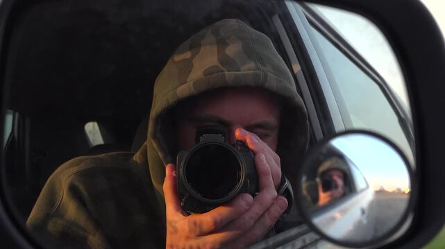 A close up view of a videographer wearing a camouflage hoodie, holding a camera up to film a self portrait reflection in a car side mirror while parked outdoors.