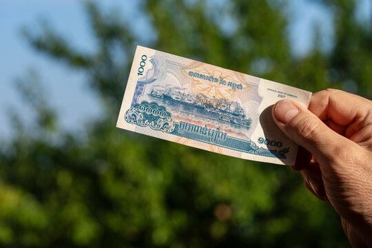 Male hand holding Cambodian riel banknote outdoors. Closeup of person holding 1000 Cambodian riel cash bill issued by National Bank of Cambodia against blurred green trees backdrop