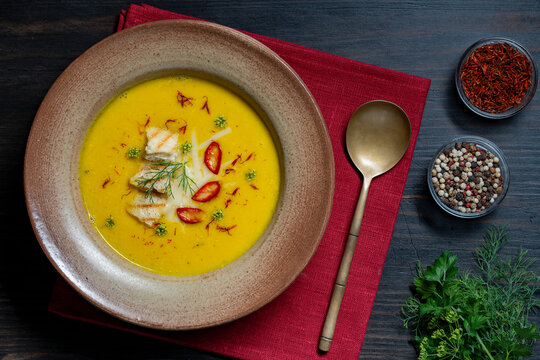 Top view of yellow pumpkin or squash cream soup, closeup. Flat lay of puree soup garnished with croutons chili pepper and saffron inside ceramic bowl on red napkin cozy restaurant culinary background