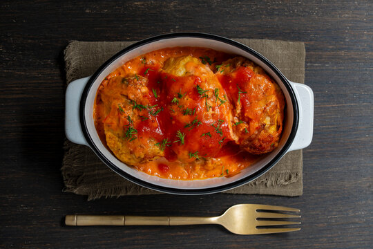 Top view of homemade cabbage rolls in ceramic baking dish. Traditional stuffed cabbage rolls or golubtsi with tomato cream sauce and fresh dill inside white casserole pan with fork on table, close up