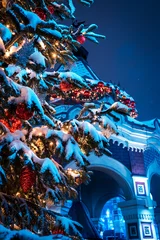 Snow Covered Christmas Tree Decorated With Red Baubles and Lights Beside Festive Building At Night © splendens