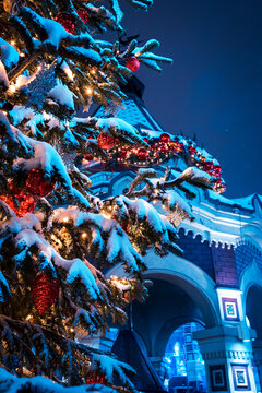 Snow Covered Christmas Tree Decorated With Red Baubles and Lights Beside Festive Building At Night