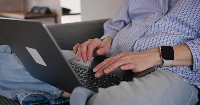 Close-up of woman typing on laptop while seated on sofa in home interior. Freelance work in casual setting.