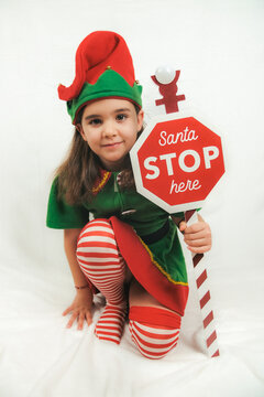 A girl dressed as one of Santa Claus's elves, posing with a sign asking Santa to stop here.