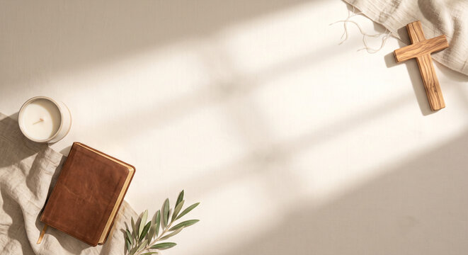 Top down view of a wooden cross, leather Bible, lit candle, and olive branch on a sunlit surface with copy space