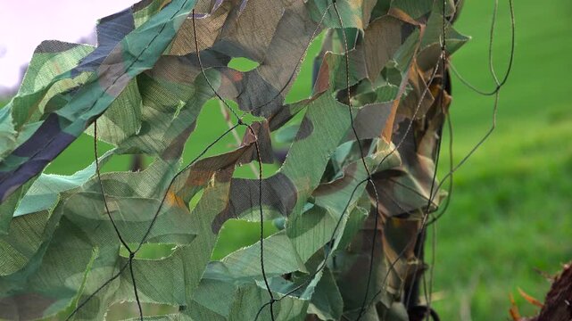 A close up of a leaf-cut camouflage mesh netting on a ground blind with wild turkey hens walking past out of focus in a vibrant green spring farm field.