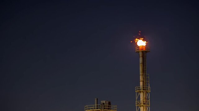 Refinery flare tower burns with bright orange flame beneath dark sky, marking active industrial gas combustion. Tall flare stack continues flickering above metal platform as heat glow expands around