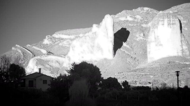 A monochrome infrared timelapse shows shifting shadows on giant rocky cliffs and a small rural house nestled in the Spanish countryside