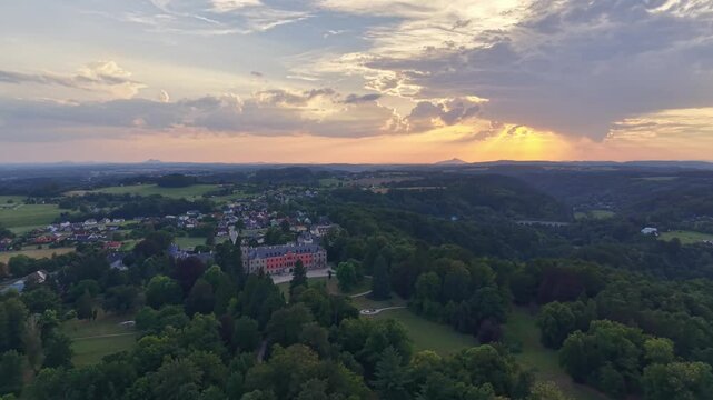 Golden hour aerial panorama of rural valley village with rolling fields, tree forests and distant hills, featuring red roof houses and stone bridge under warm sunset light, long shadows and sky