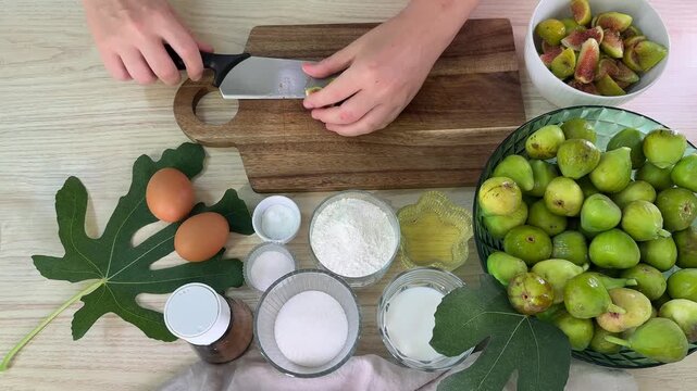 a person handles green figs and ingredients including eggs, flour, and sugar on a light wooden surface with fig leaves